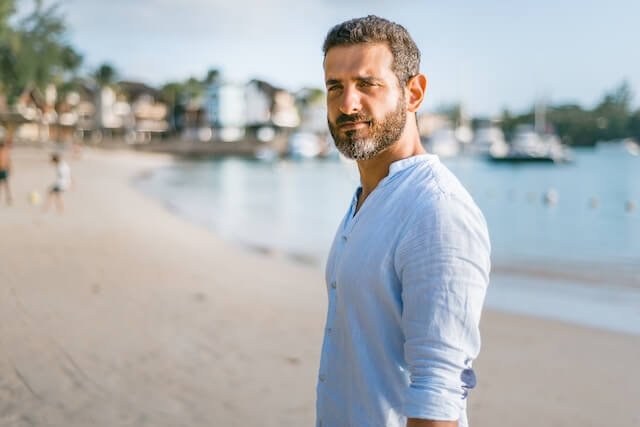 Photo of the side profile of a man with grey and black hair on the beach in a light blue shirt.